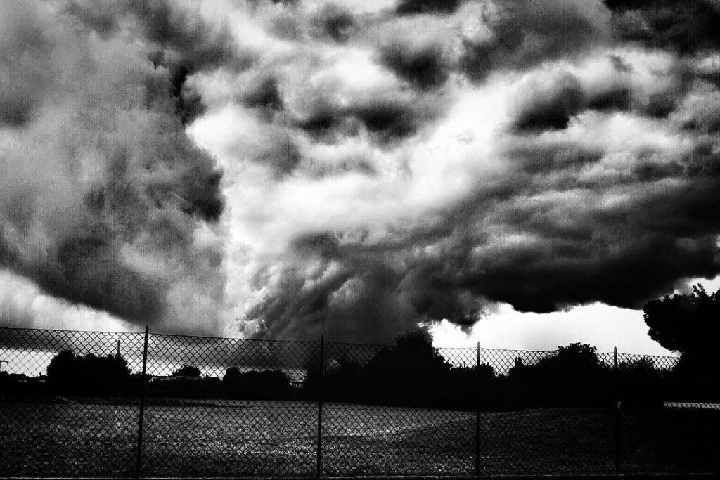 Tempesta in arrivo con nuvole scure sopra una rete metallica, fotografia in bianco e nero ad alto contrasto che simboleggia la fragilità umana contro la natura.