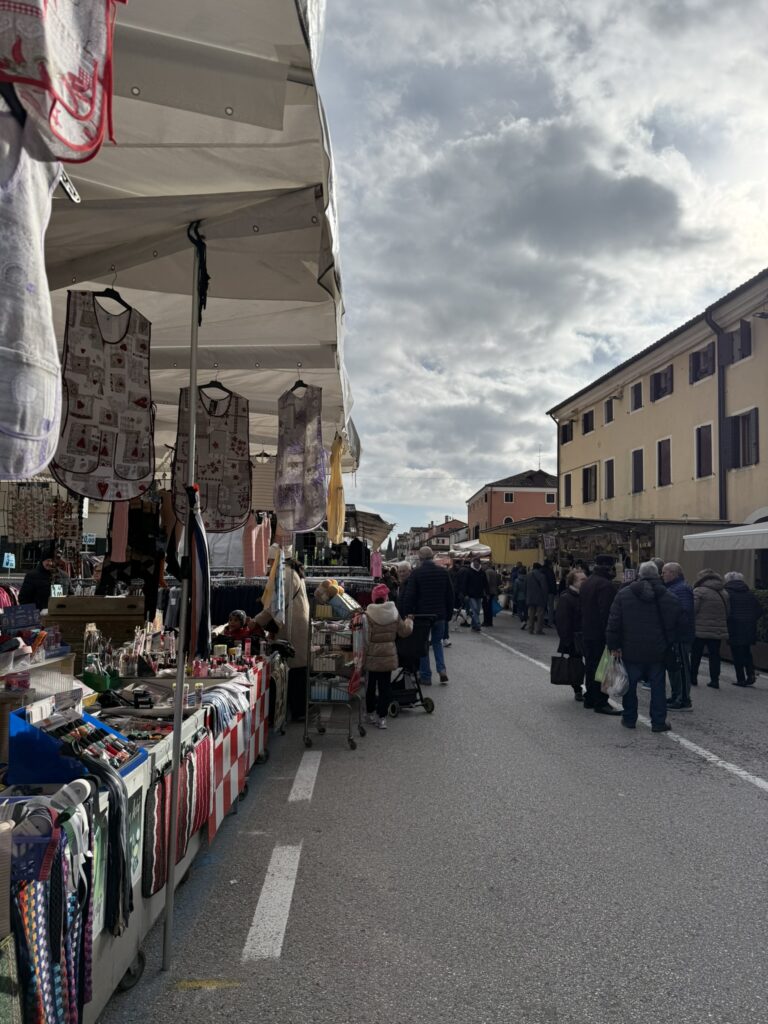 Mercato settimanale di Mirano sotto tende bianche sventolanti, banchi di abbigliamento e oggetti colorati, folla di persone in cappotti contro cielo grigio nuvoloso, edifici veneti sullo sfondo – vita autentica, anti-algoritmo.