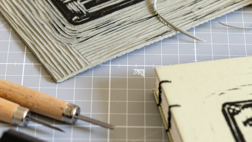 Strumenti di legatoria artigianale sul piano da lavoro del laboratorio Saimai: coltelli, filo e taccuino cucito a mano con copertina in carta da upcycling