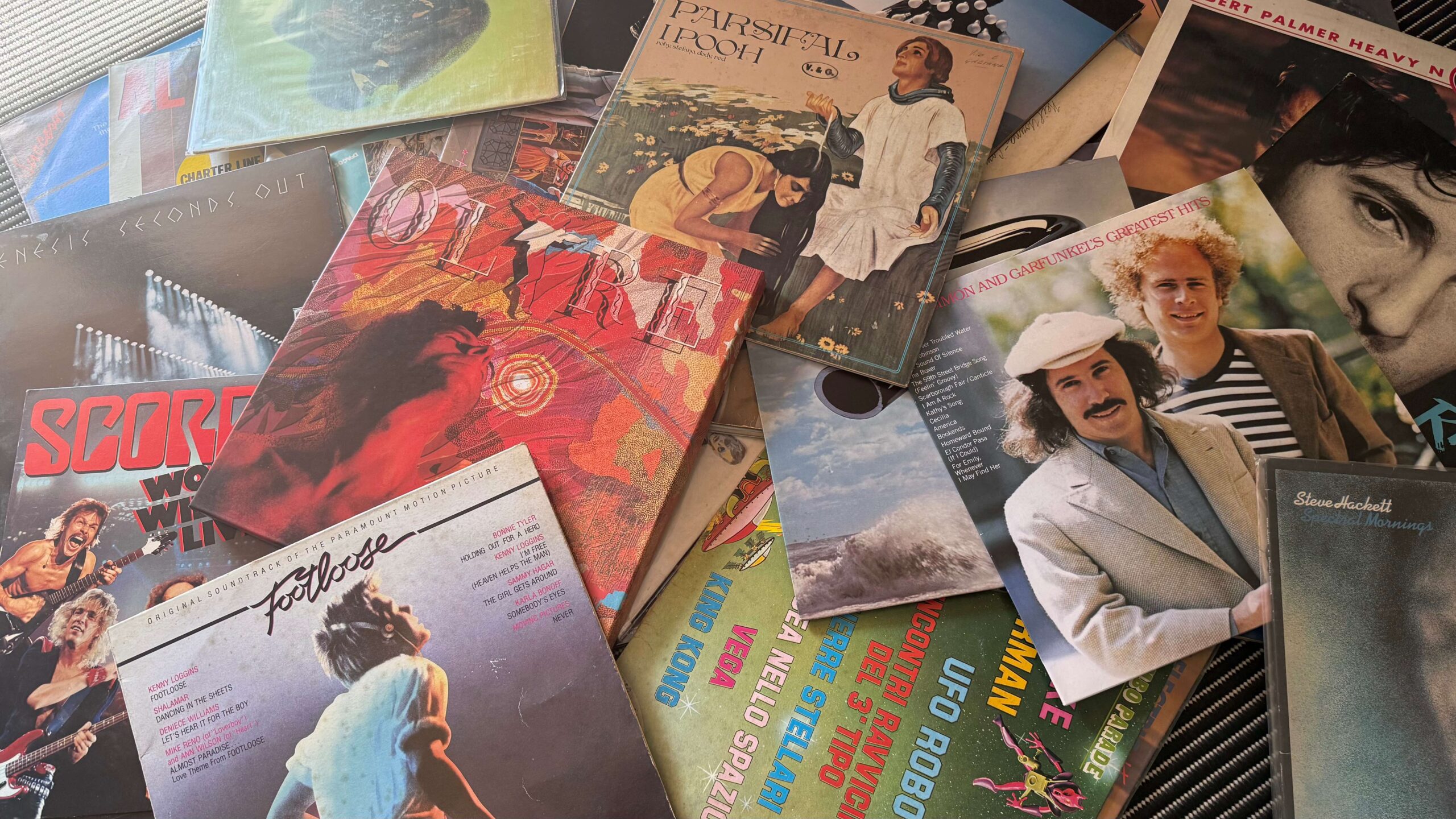 Una collezione di dischi in vinile sparsi come frammenti di una vita musicale: Genesis, Scorpions, Footloose, I Pooh, Simon & Garfunkel, Steve Hackett. Plastica nera e carta stampata. comprare vinili nel 2026 contro l'algoritmo.