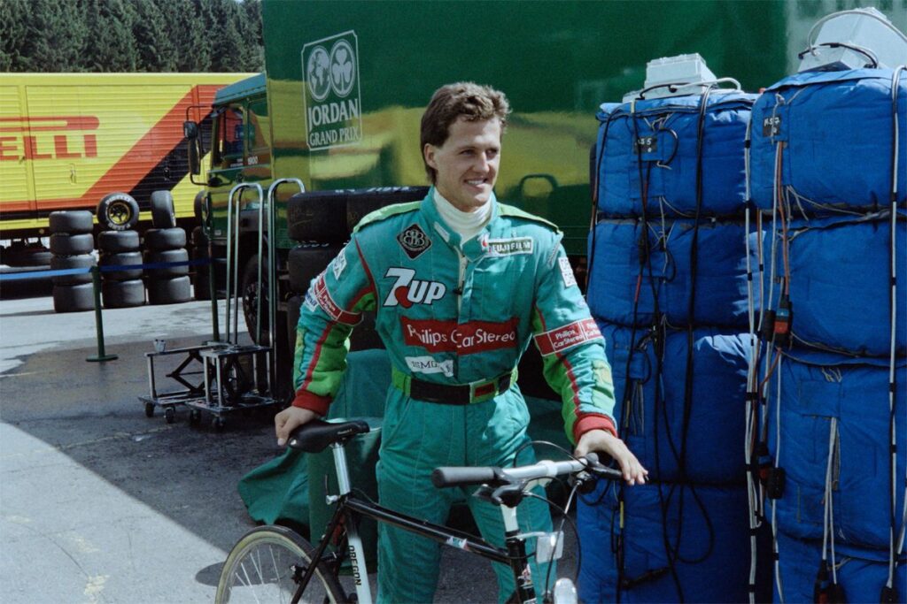 Un giovane Michael Schumacher, a ventidue anni, posa con la tuta ignifuga verde della Jordan 191 nel paddock di Spa-Francorchamps, con un'espressione decisa e le braccia incrociate.